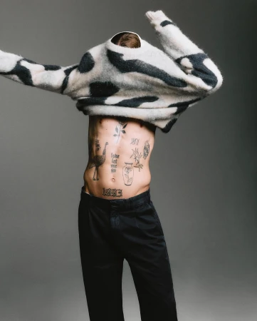 Shirtless man with torso tattoos pulling off a patterned monochrome sweater against a grey studio backdrop.