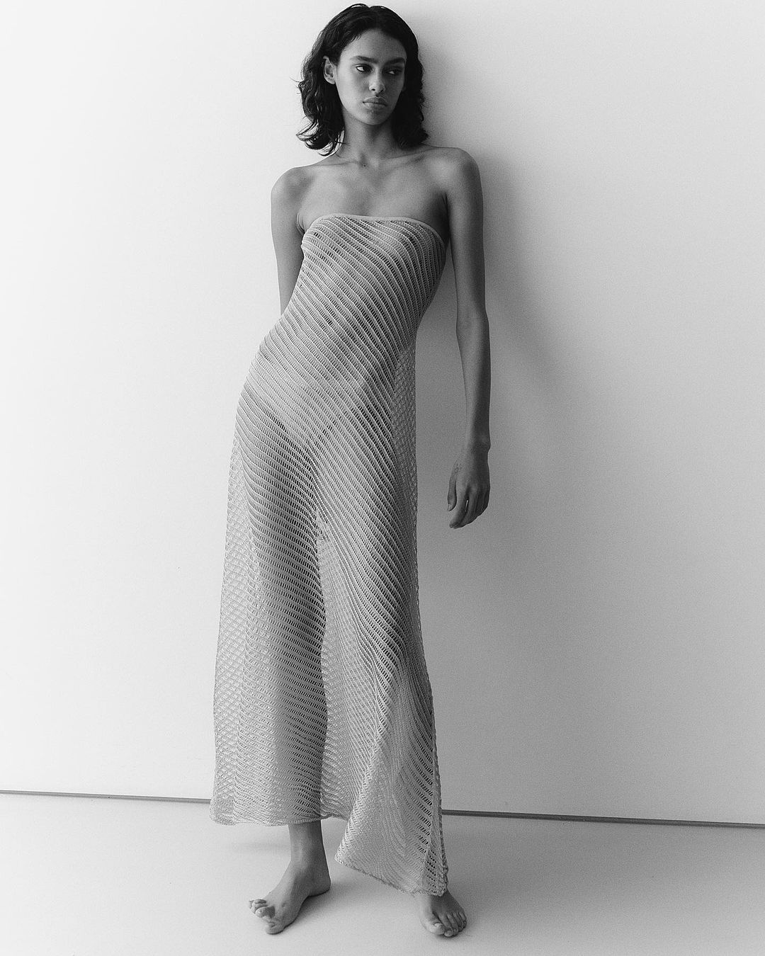 Monochrome portrait of a woman wearing a sheer ribbed strapless dress standing against a plain light wall