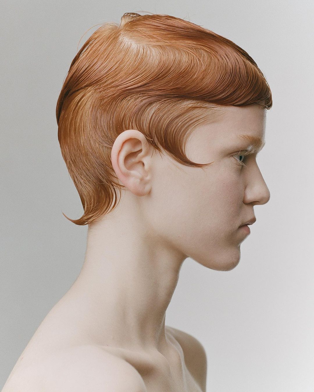 Portrait of a fair-skinned adult woman with an expressive profile, striking copper hair styled in retro waves, and a light gray background.