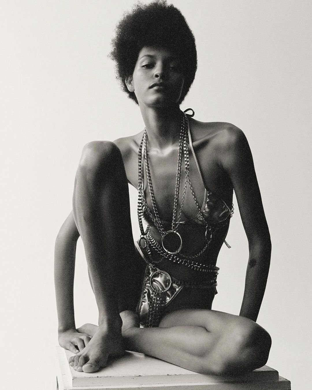 A model poses in a studio wearing a metallic bikini and layered chain jewelry, with a neutral backdrop and soft, diffused lighting.