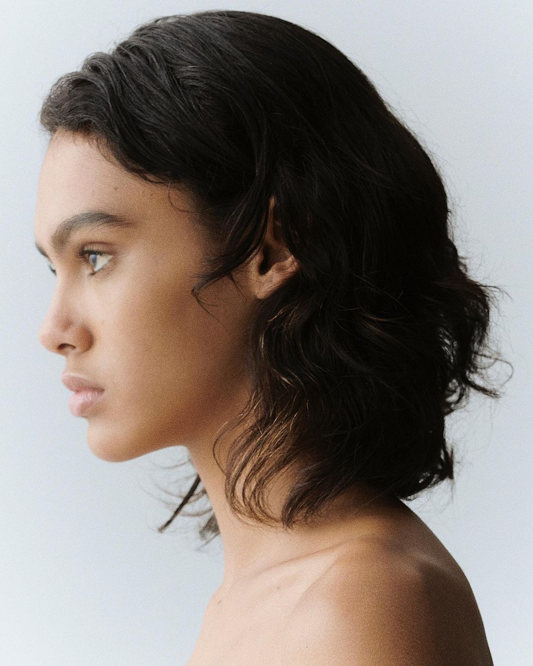 Profile portrait of a young woman with dark wavy hair against a plain light background photographed by Anna Daki