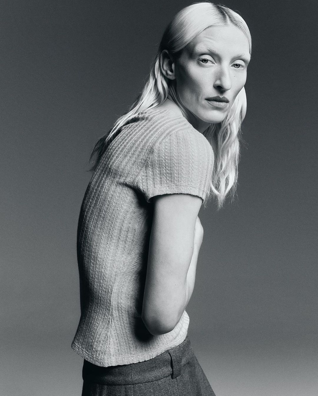 Monochrome studio portrait of a woman with pale skin and blonde hair, wearing a short-sleeved knitted shirt and pants, side view.