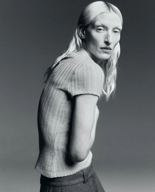 Monochrome studio portrait of a woman with pale skin and blonde hair, wearing a short-sleeved knitted shirt and pants, side view.