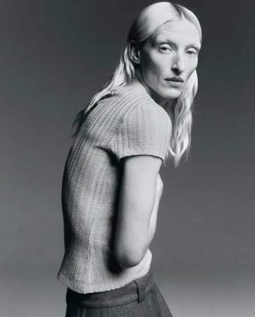 Monochrome studio portrait of a woman with pale skin and blonde hair, wearing a short-sleeved knitted shirt and pants, side view.