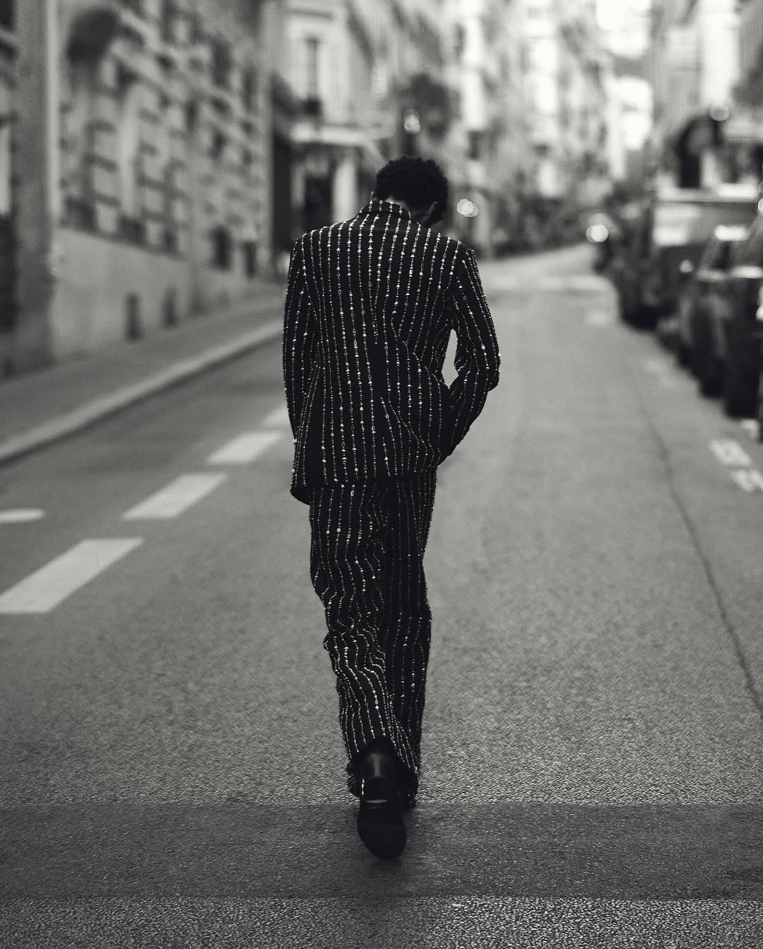A person seen from behind walking away down a narrow Parisian street wearing a pinstripe suit.