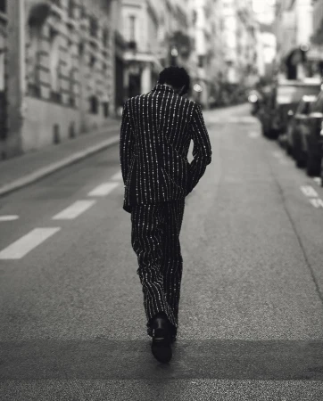 A person seen from behind walking away down a narrow Parisian street wearing a pinstripe suit.