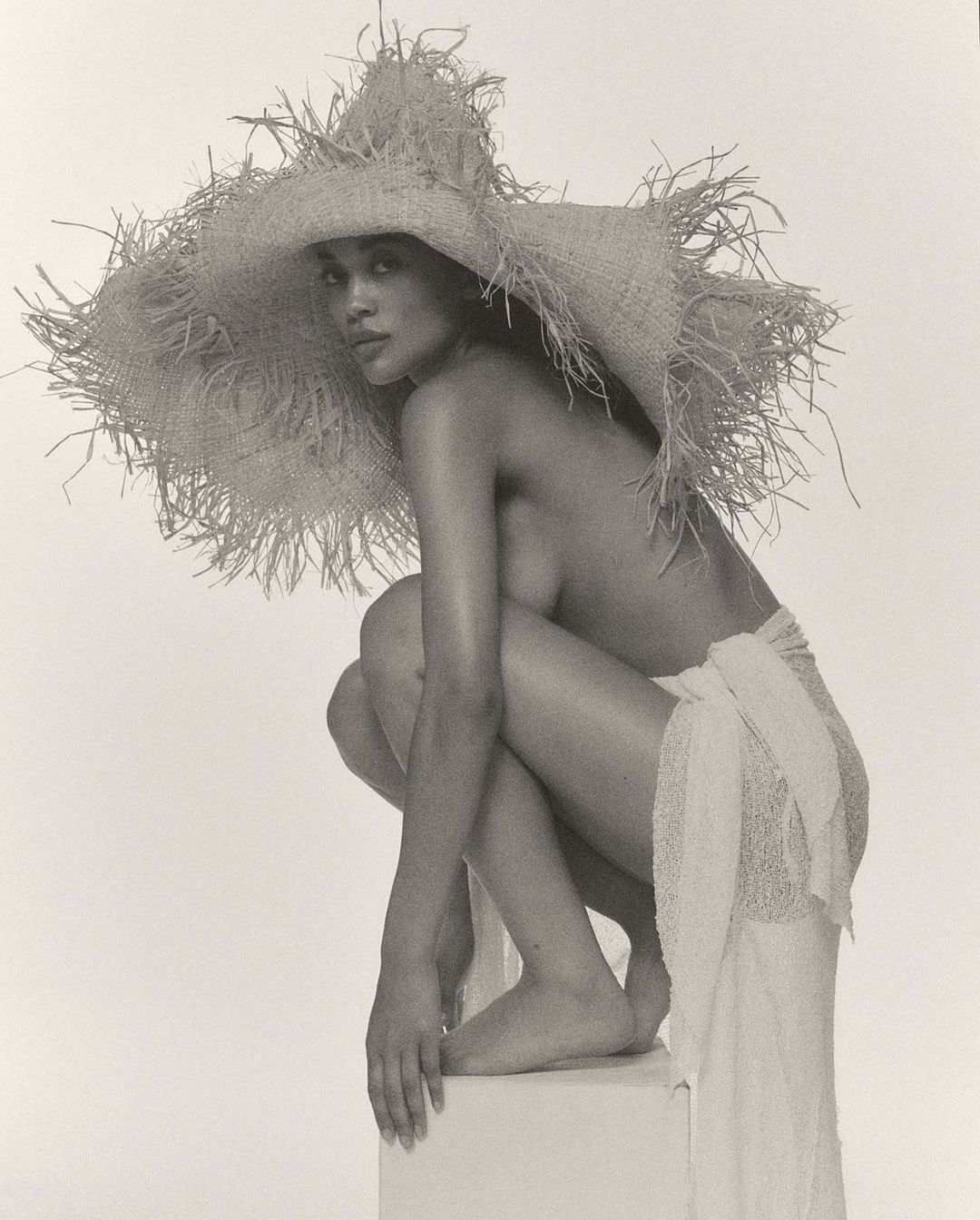 Striking black and white studio portrait of a topless woman with a large straw hat, crouching on a white block, draped in white fabric.
