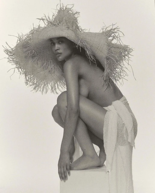 Striking black and white studio portrait of a topless woman with a large straw hat, crouching on a white block, draped in white fabric.