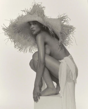 Striking black and white studio portrait of a topless woman with a large straw hat, crouching on a white block, draped in white fabric.