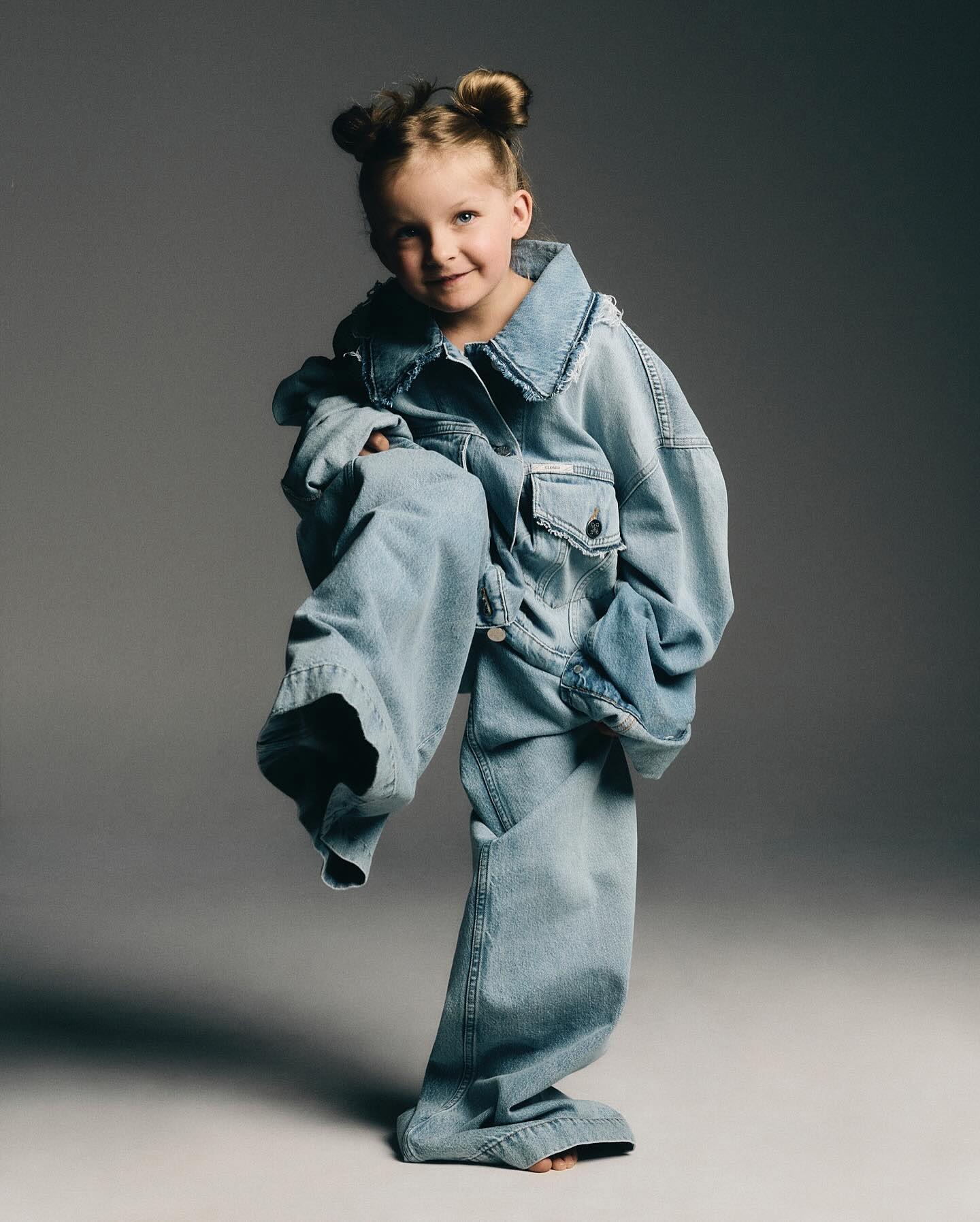 Young child posing in an oversized denim jacket and jeans in a studio setting with gray background and neutral, diffused lighting.