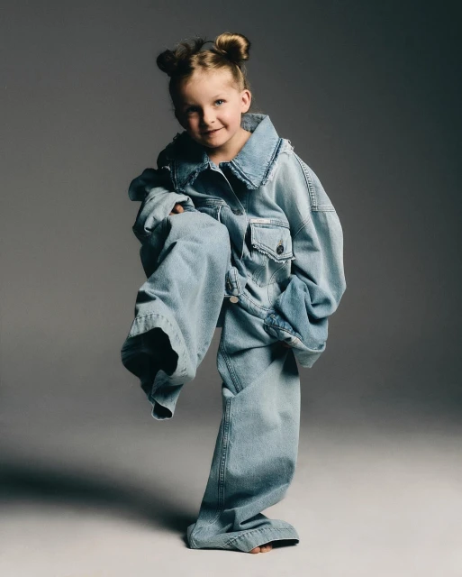 Young child posing in an oversized denim jacket and jeans in a studio setting with gray background and neutral, diffused lighting.