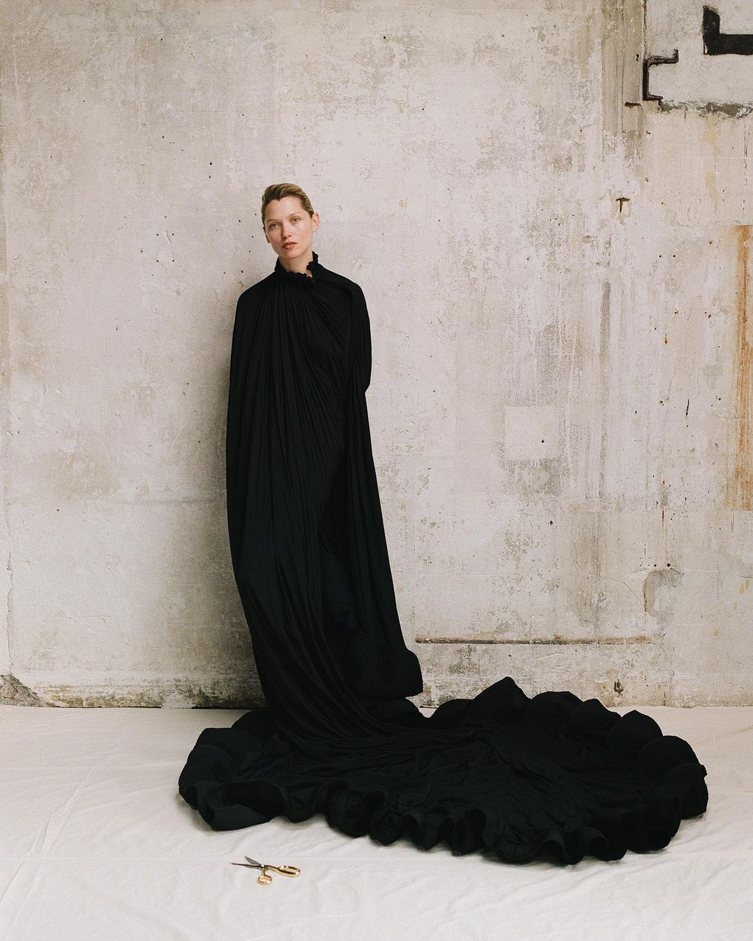 Woman in black gown with a long train standing in front of a concrete wall, with a pair of gold scissors on the floor.