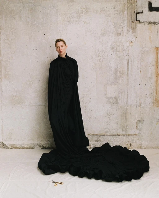 Woman in black gown with a long train standing in front of a concrete wall, with a pair of gold scissors on the floor.