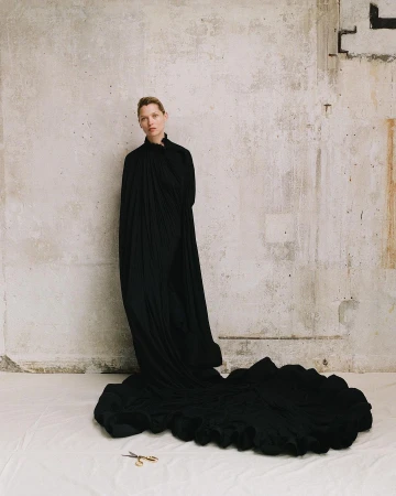 Woman in black gown with a long train standing in front of a concrete wall, with a pair of gold scissors on the floor.