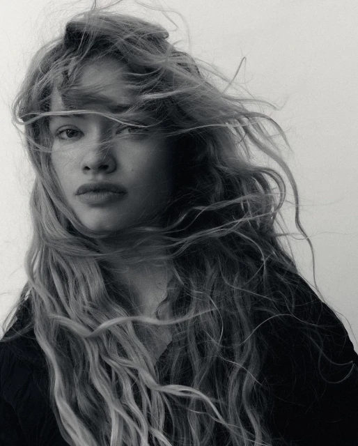 Black and white portrait of a young adult female with long wavy hair blowing in the wind against a neutral background. Beauty shot.