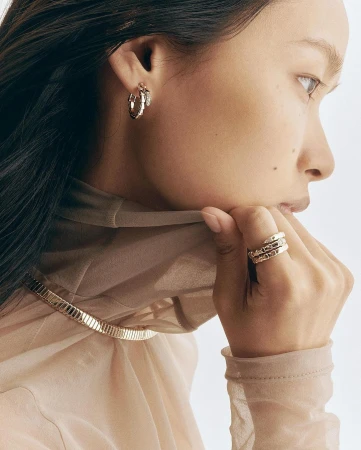 Close-up portrait of a woman wearing gold hoop earrings, layered rings, a rigid choker necklace, and a sheer beige top.