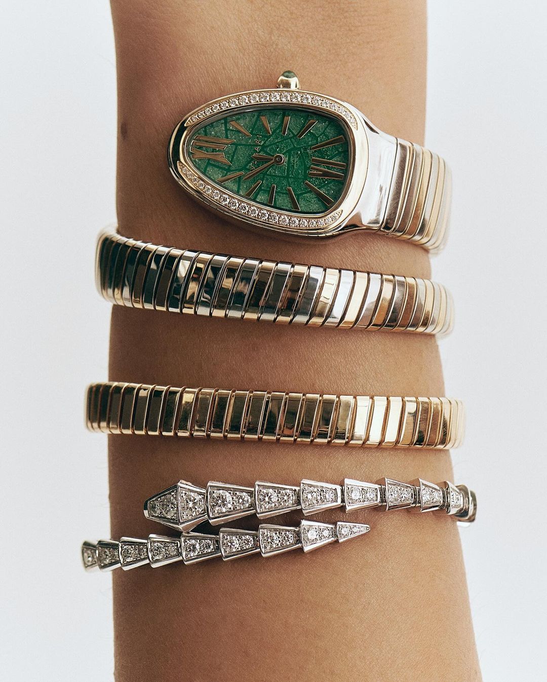 Wrist adorned with a gold and silver snake-shaped watch with a green face, complemented by three flexible bangle bracelets against a white backdrop.
