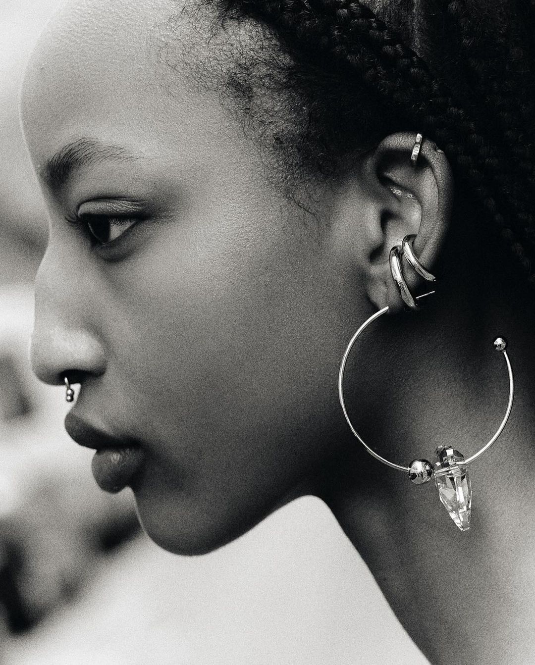 Profile view of an adult female with dark hair in braids, wearing various piercings and a large hoop earring with a crystal accent.