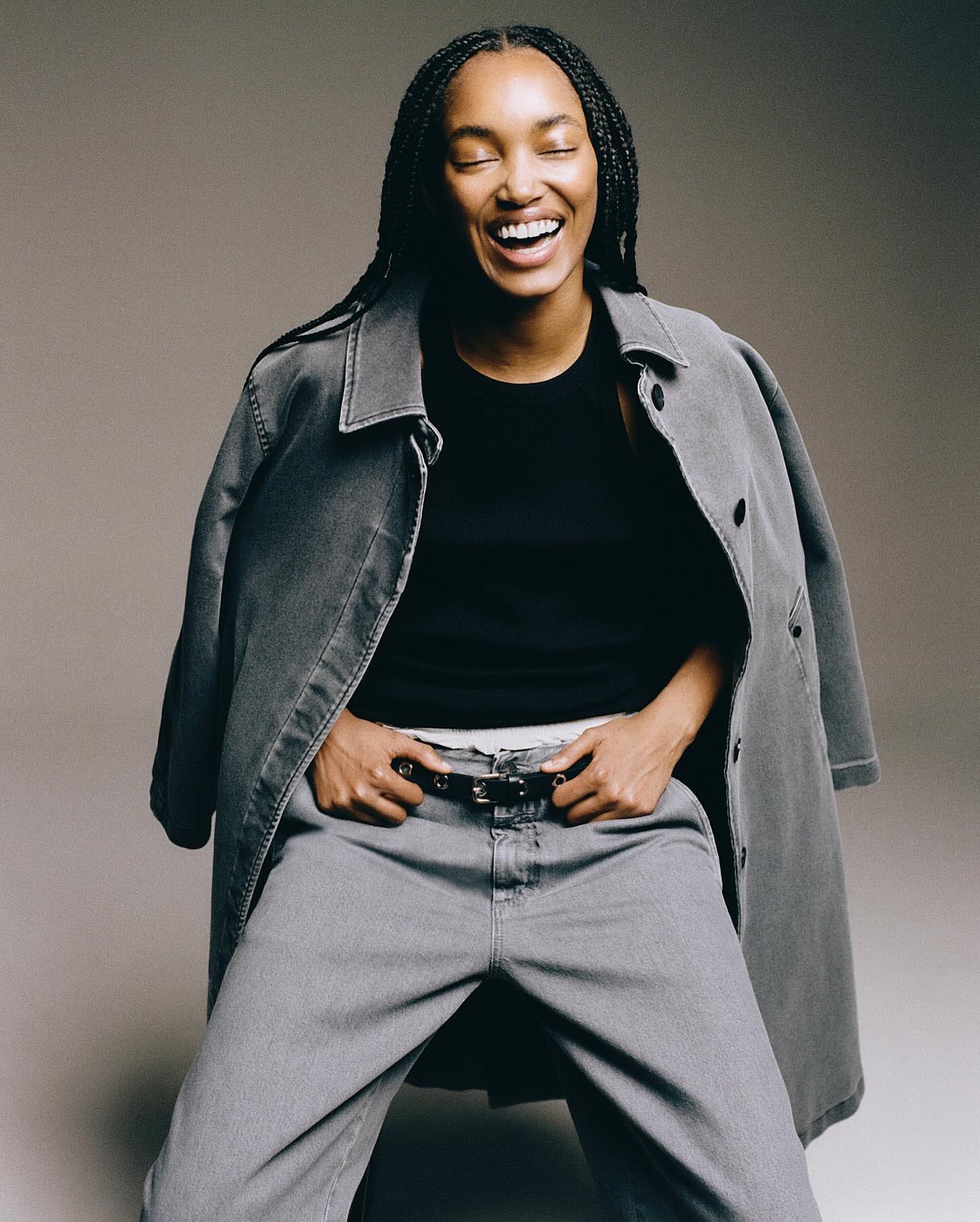 Stylish black woman with braided hair laughing in a studio shot. She wears grey jeans and jacket with a black top in a portrait.