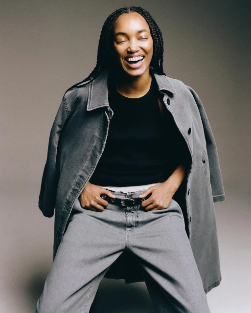 Stylish black woman with braided hair laughing in a studio shot. She wears grey jeans and jacket with a black top in a portrait.