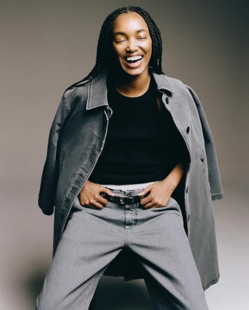 Stylish black woman with braided hair laughing in a studio shot. She wears grey jeans and jacket with a black top in a portrait.