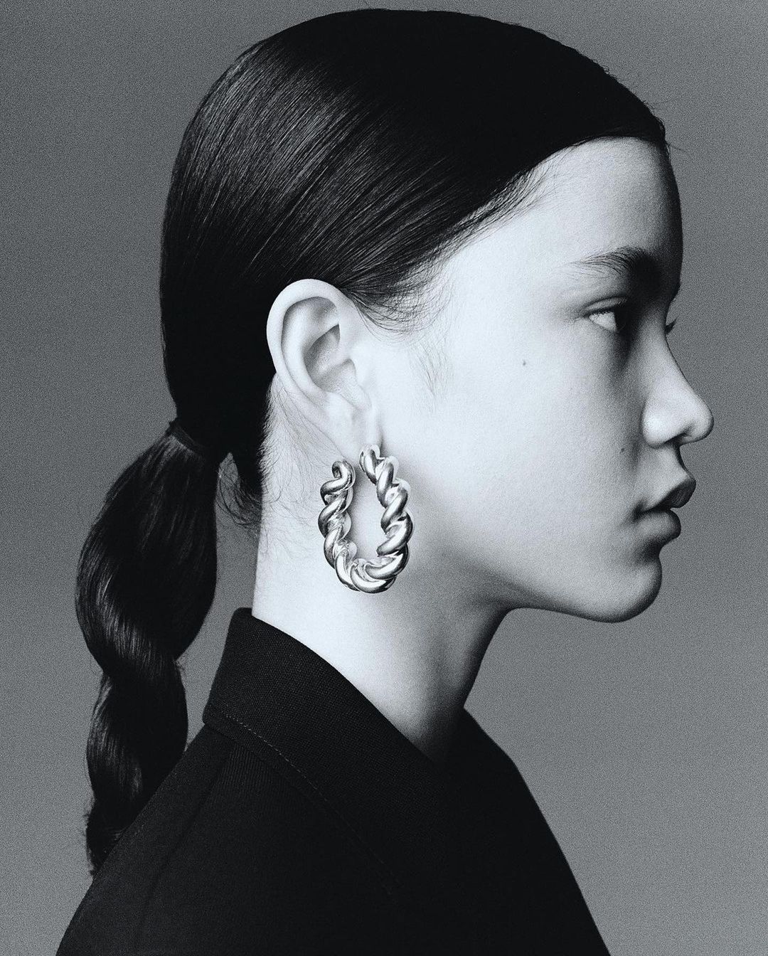 Profile portrait of an adult female with braided hair wearing hoop earrings and a dark top against a grey background. Studio shot.