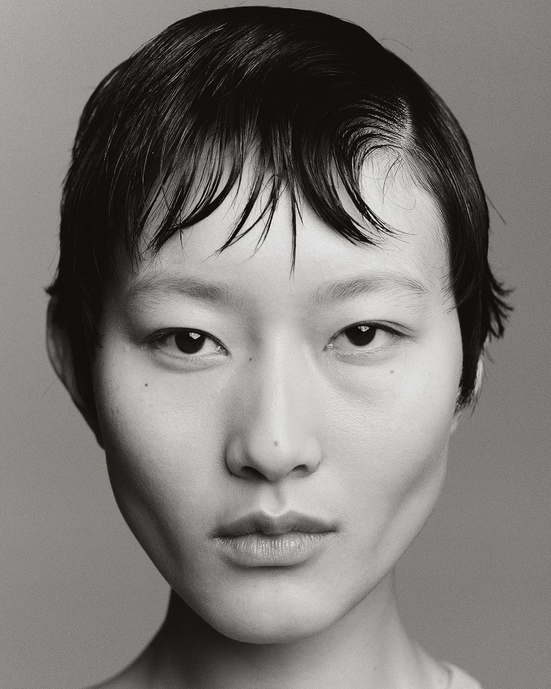 Close-up black and white portrait of an Asian female with short dark hair and natural makeup, set against a plain grey background.