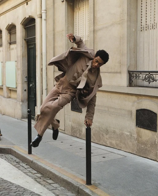 An adult man in a tan suit leaps from a metal pole on a city street, his body suspended mid-air with a background of tall buildings.