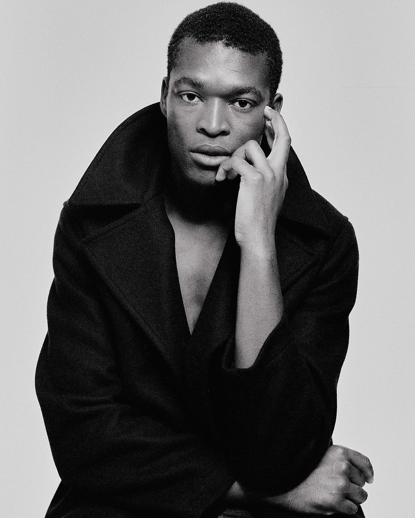 Monochrome portrait of a dark-skinned male in a black coat, with hand on his face, set against a neutral grey backdrop in a studio setting.