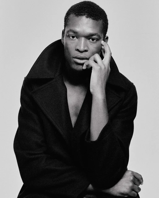 Monochrome portrait of a dark-skinned male in a black coat, with hand on his face, set against a neutral grey backdrop in a studio setting.