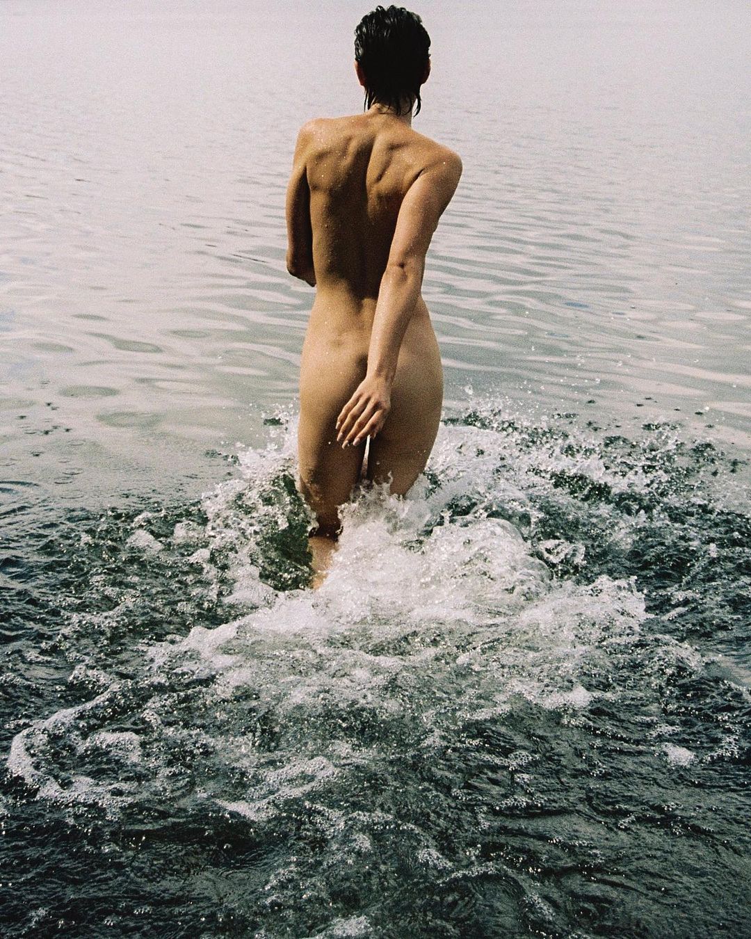 A lone woman walks into a misty body of water, her back facing the camera, creating ripples and splashes on the lake's surface.