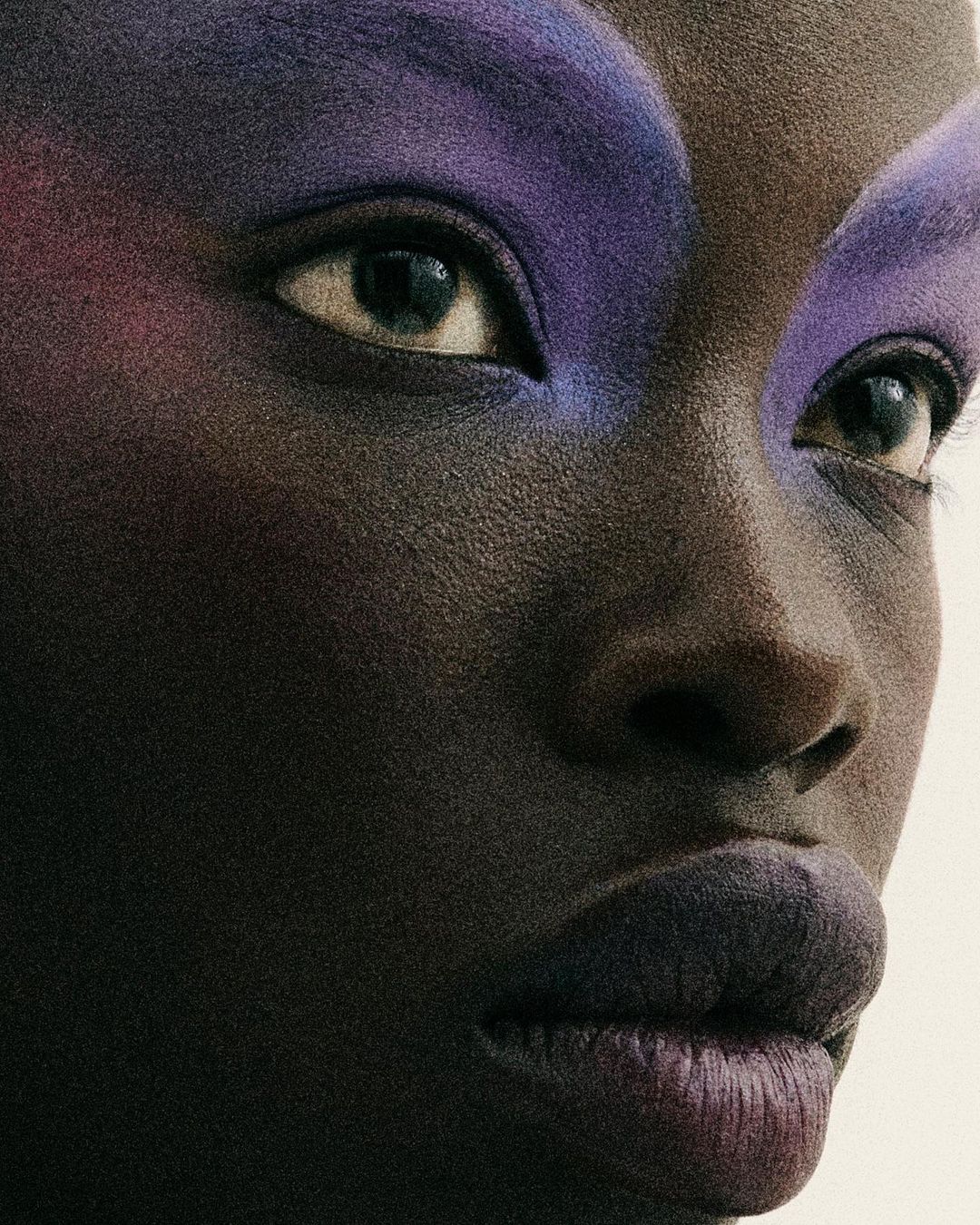 Close-up portrait of a Black woman's face with dramatic purple makeup on her eyelids and lips in a beauty editorial style against a neutral background.
