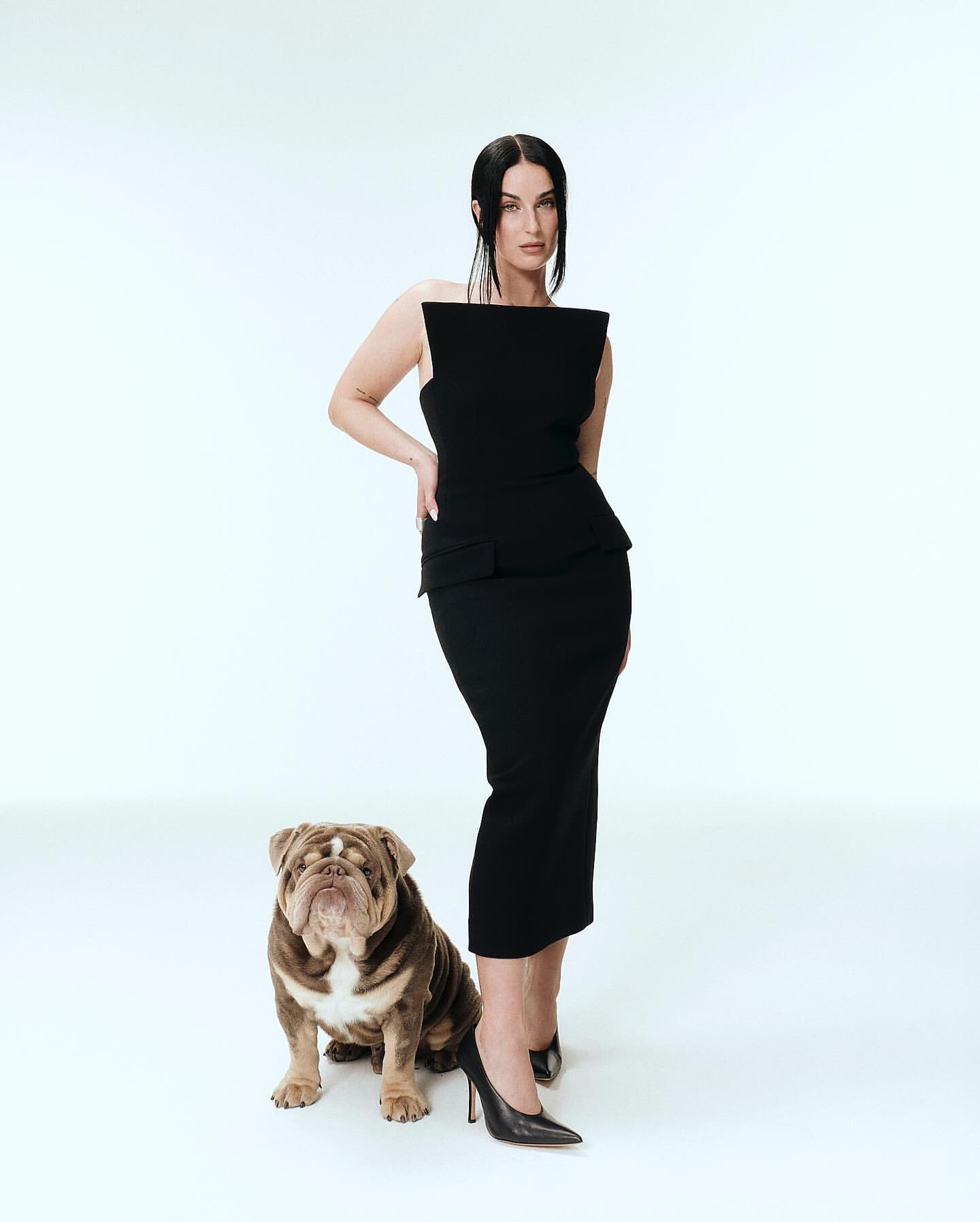 Elegant woman in a black dress poses in a white studio with her bulldog, creating a classic, fashionable, and polished portrait.