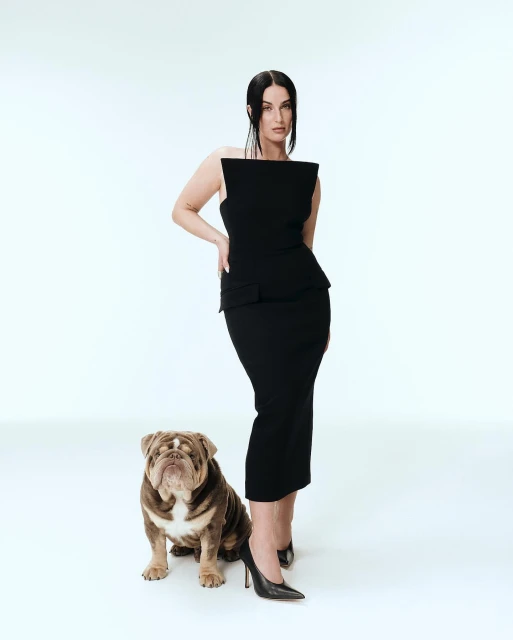 Elegant woman in a black dress poses in a white studio with her bulldog, creating a classic, fashionable, and polished portrait.