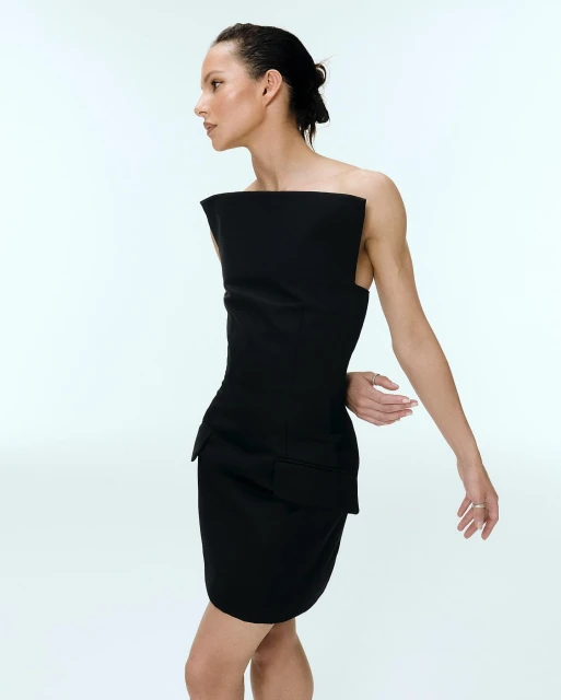 A woman poses in a stylish black dress against a white backdrop, highlighting the dress's unique structural design and the model's elegant pose.