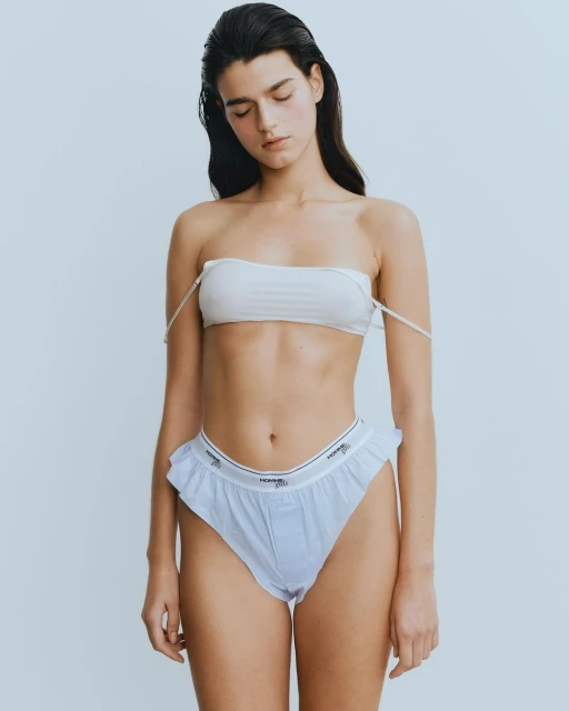 A woman poses indoors wearing a white bandeau top and white underwear. The image features soft lighting and a minimalist aesthetic.