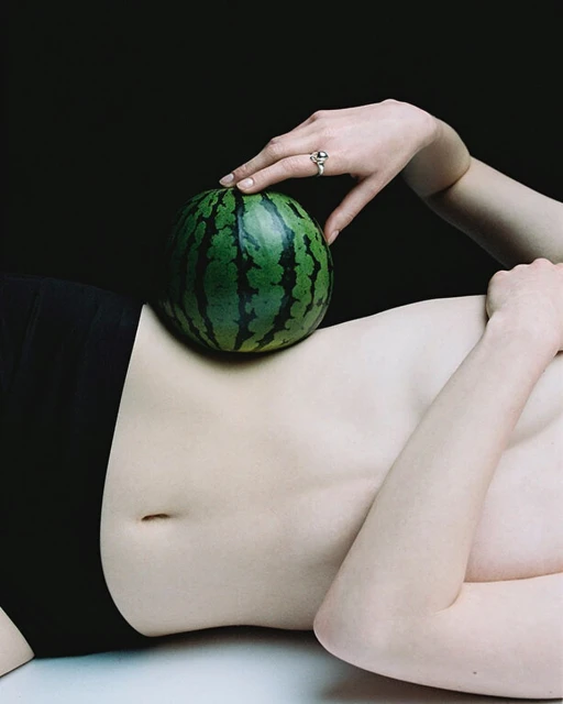An adult female figure laying down, supporting a watermelon on their torso, hand on top of the watermelon. Black background, studio lighting.