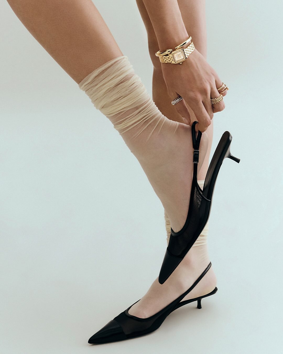 Close-up of a woman putting on a black slingback heel with a sheer tan sock, accessorized with rings and a gold watch, in a studio setting.