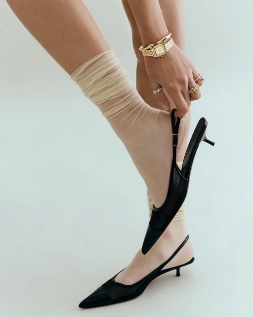 Close-up of a woman putting on a black slingback heel with a sheer tan sock, accessorized with rings and a gold watch, in a studio setting.