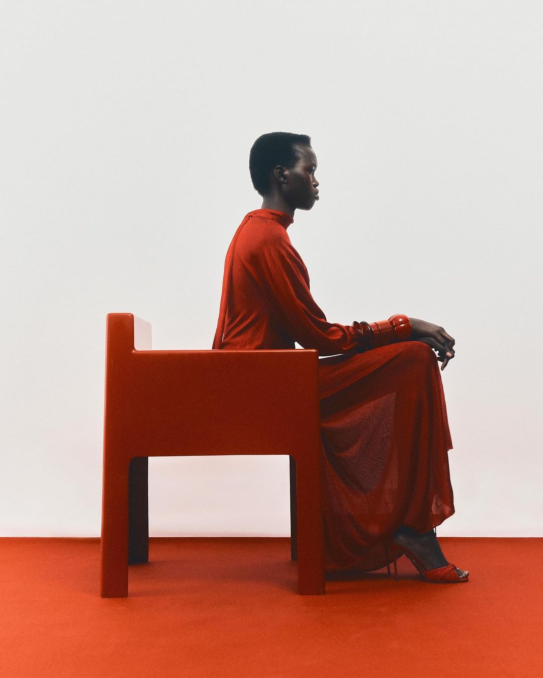 A Black woman wearing a red dress seated on a matching red chair against a plain white background.