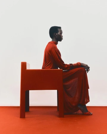 A Black woman wearing a red dress seated on a matching red chair against a plain white background.