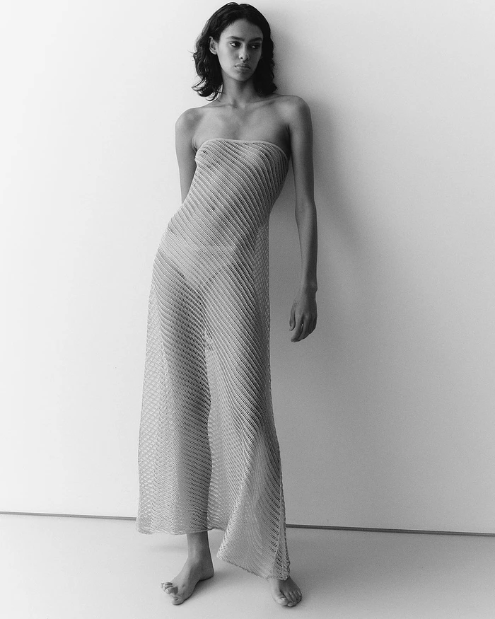 Monochrome portrait of a woman wearing a sheer ribbed strapless dress standing against a plain light wall