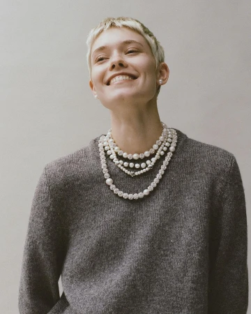 Smiling model with short blonde hair wearing a grey knit sweater and layered pearl necklaces photographed in Paris by Anna Daki