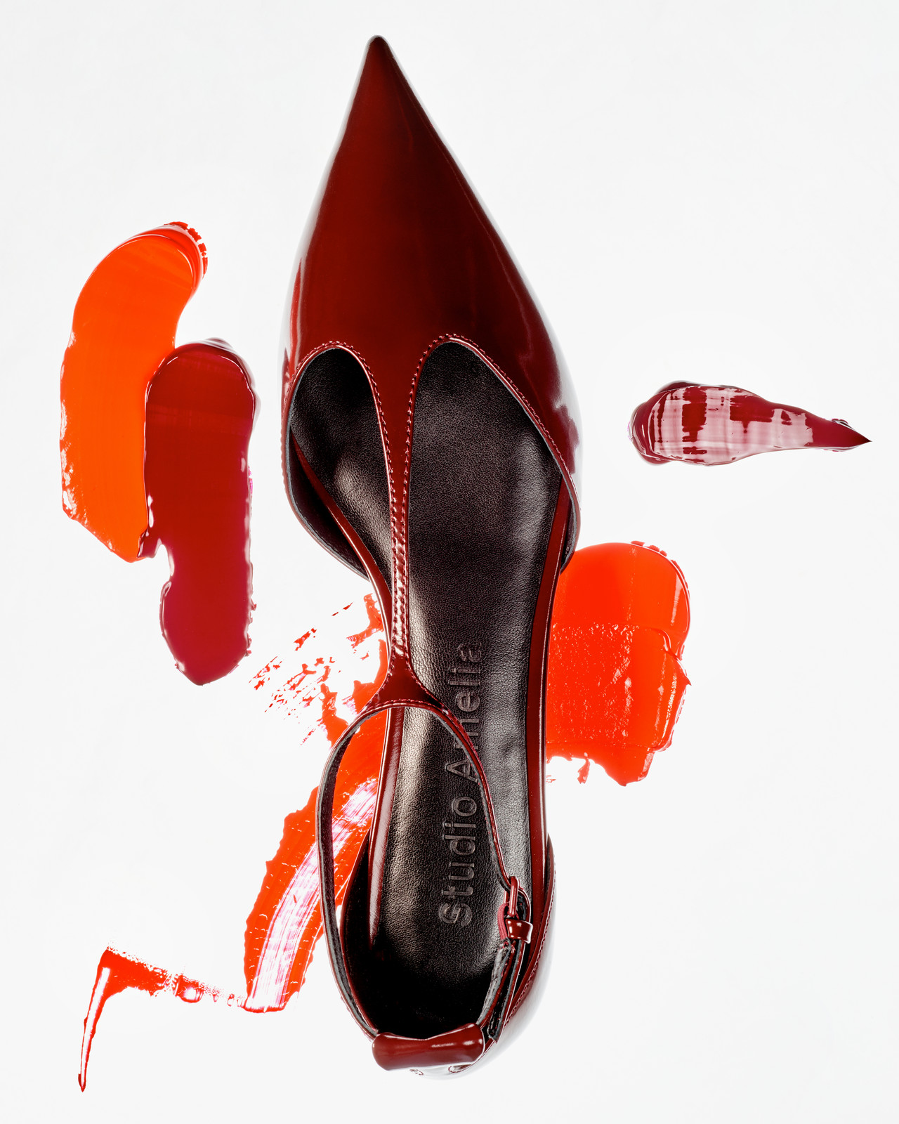 Overhead shot of a glossy, dark red patent leather slingback heel surrounded by strokes of lipstick and lipgloss against a white backdrop.
