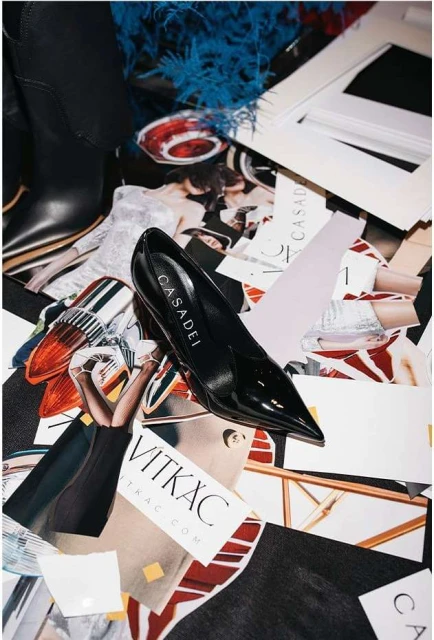 Still life of a glossy black Casadei stiletto heel atop fashion magazine and brand paper cutouts, capturing a high-fashion editorial aesthetic.