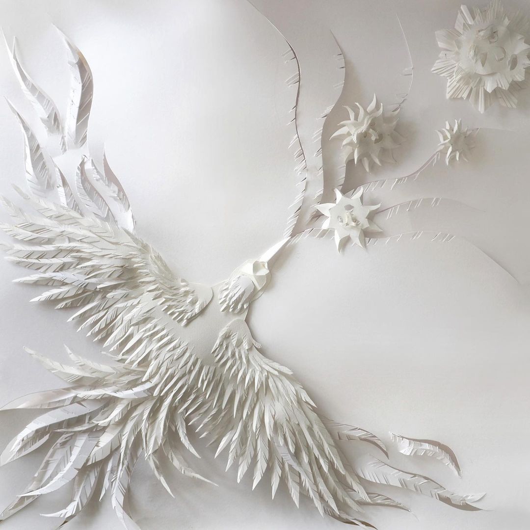 Elaborate paper art sculpture of a phoenix bird with wing details and starburst designs, all in white, against a seamless white backdrop.