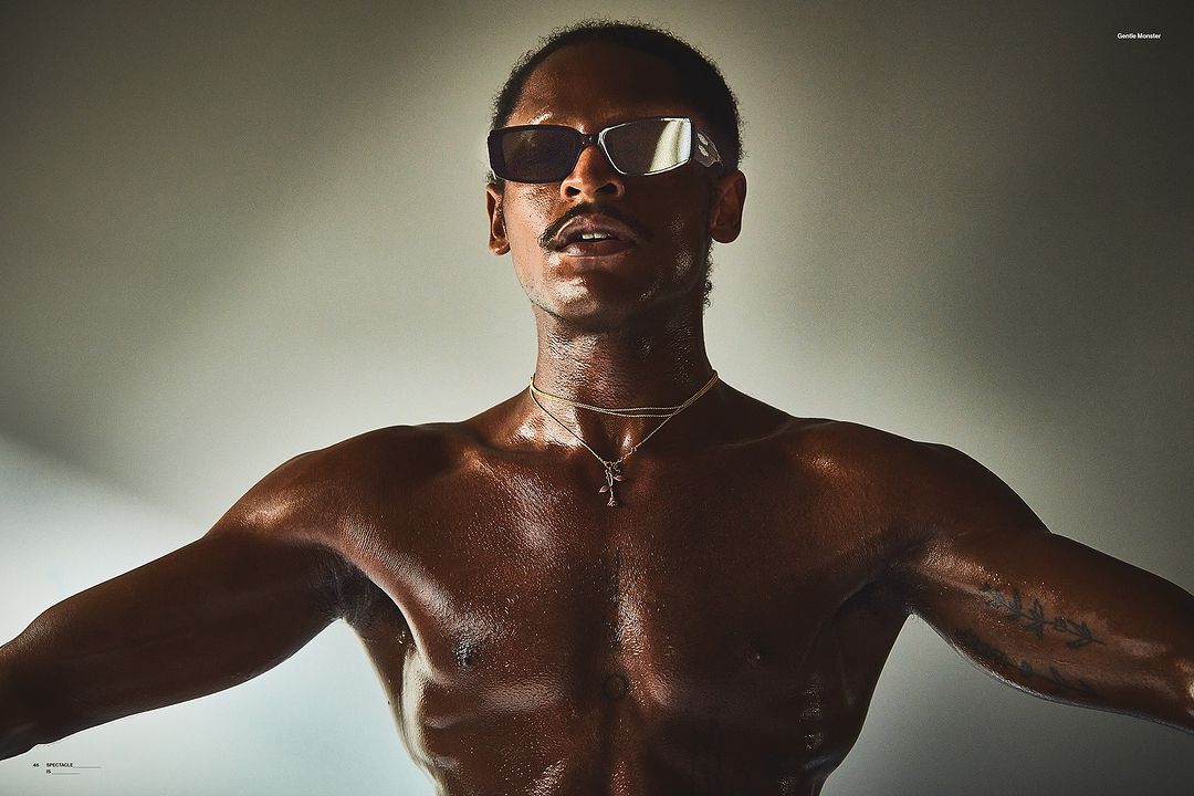 Close-up studio portrait of a dark-skinned adult male with a mustache wearing sunglasses and jewelry with visible tattoo on his arm.