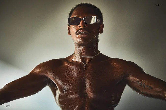 Close-up studio portrait of a dark-skinned adult male with a mustache wearing sunglasses and jewelry with visible tattoo on his arm.