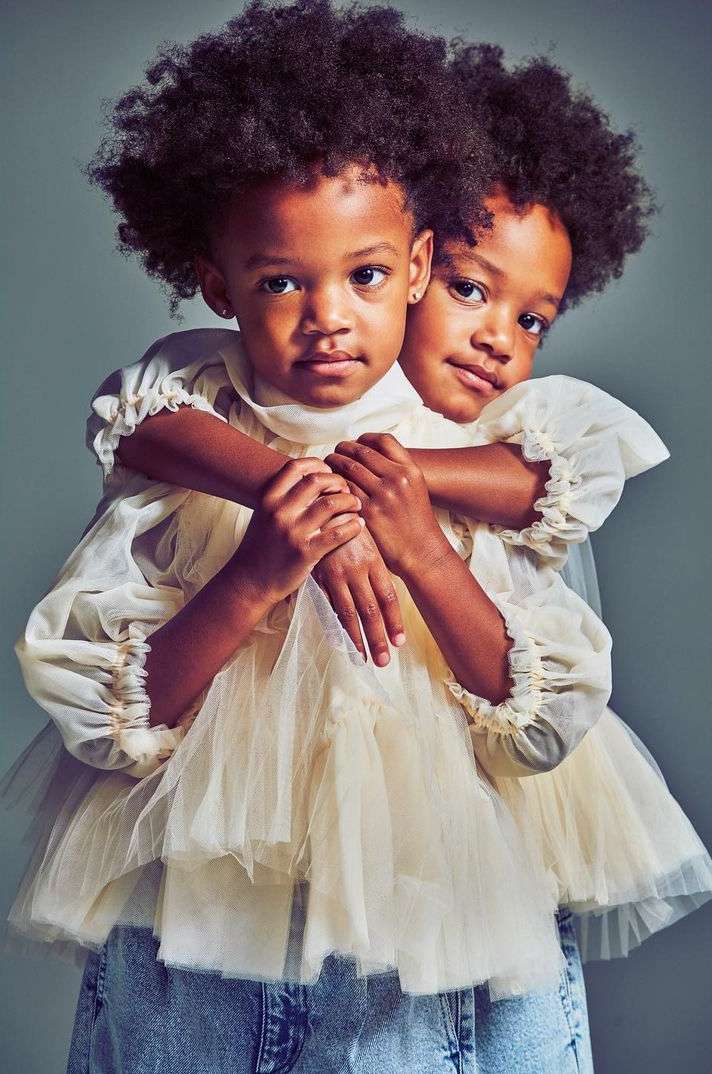 Portrait of two young African American girls embracing and wearing matching dresses and denim jeans against a neutral backdrop.