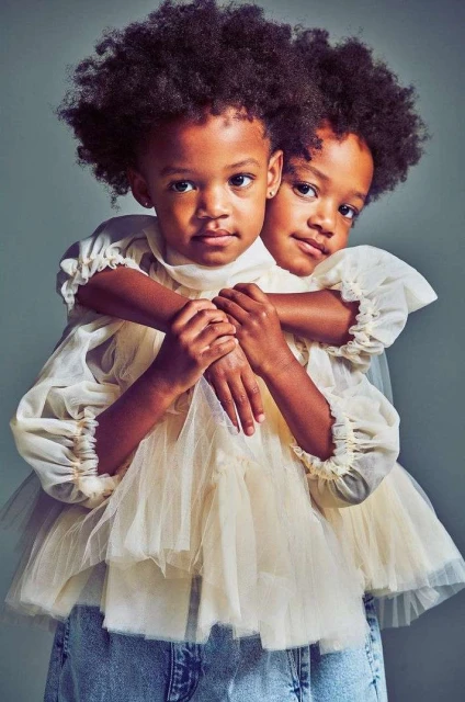 Portrait of two young African American girls embracing and wearing matching dresses and denim jeans against a neutral backdrop.